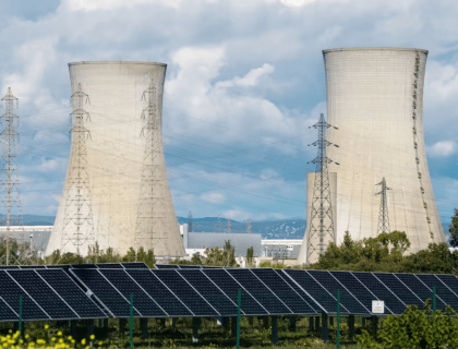 Thumbnail image for the 'Energizing Australia: Renewable Versus Nuclear in the National Debate' article, featuring a solar panel farm in the foreground with large nuclear power plant cooling towers in the background, visually representing the contrast and discussion points between renewable and nuclear energy options discussed in the summary of the Moolah show hosted by Omar de Silva.