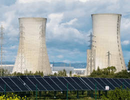 Thumbnail image for the 'Energizing Australia: Renewable Versus Nuclear in the National Debate' article, featuring a solar panel farm in the foreground with large nuclear power plant cooling towers in the background, visually representing the contrast and discussion points between renewable and nuclear energy options discussed in the summary of the Moolah show hosted by Omar de Silva.